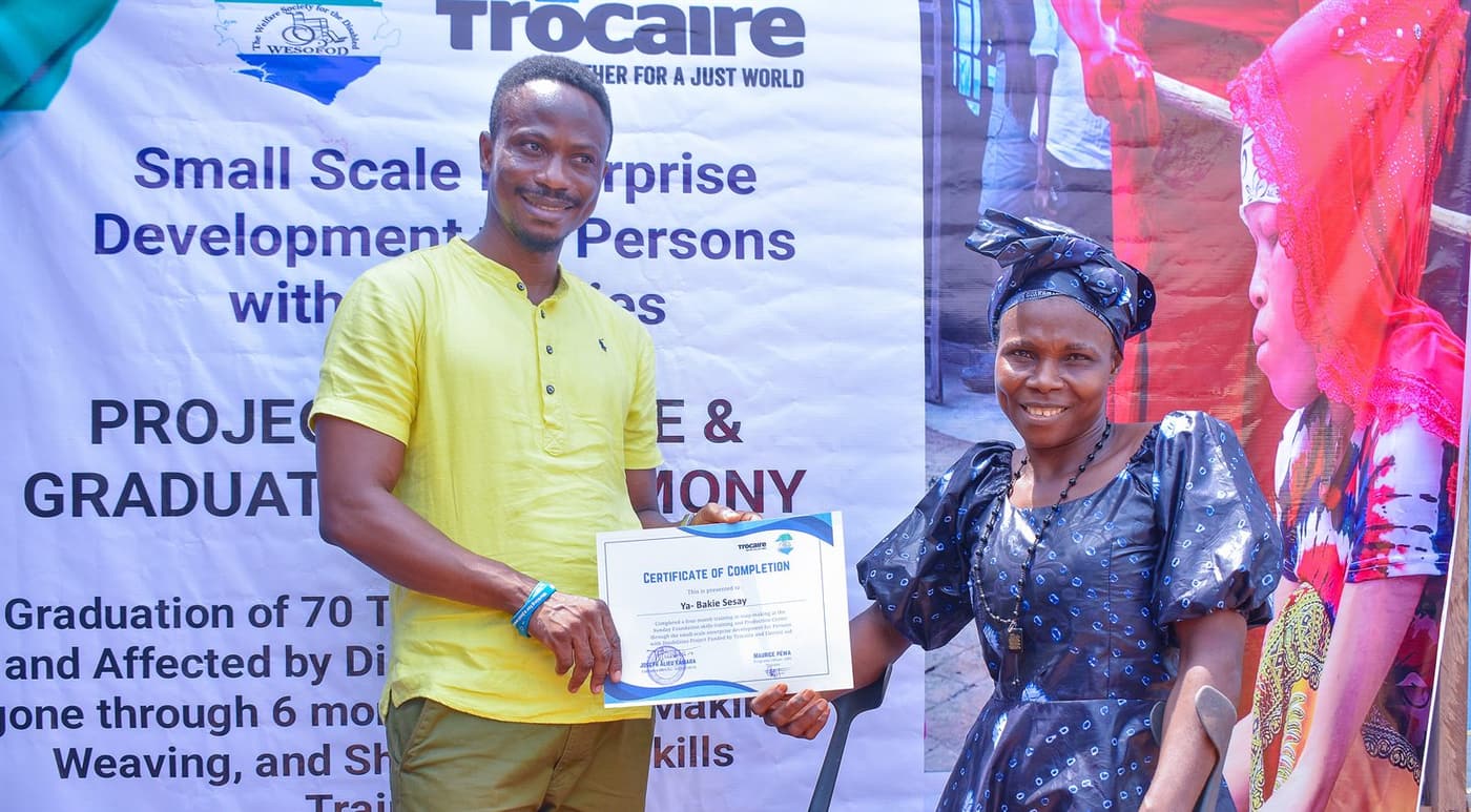 A WESOFOD staff member handing a certificate of completion to a graduate on crutches at the Small Scale Enterprise Development ceremony