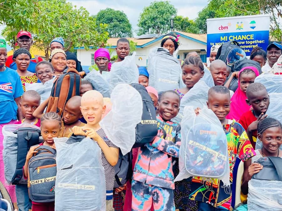 A crowd of smiling children, among them a child with albinism, hold up new school bags in front of an MI-Chance banner.