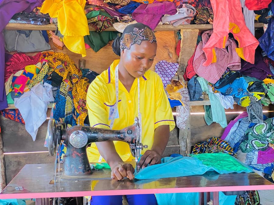 A young woman with a tape measure round her neck sews turquoise fabric on a treadle sewing machine, surrounded by finished garments.