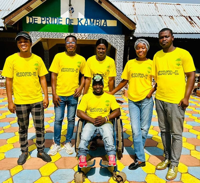WESOFOD-SL staff in yellow organization T-shirts outside the Kambia office, a colleague seated in a wheelchair at the front of the group